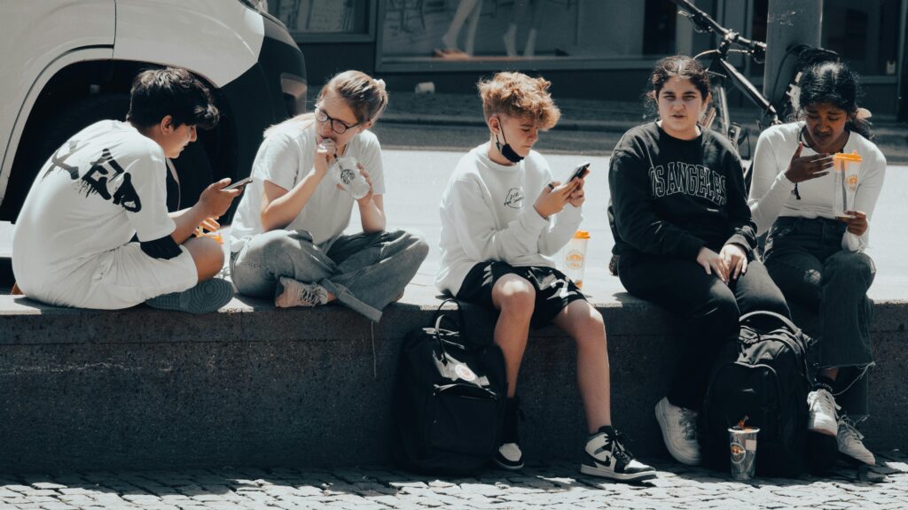 Group of teenagers sitting on a sidewalk, interacting with phones and drinks, showcasing diversity and street style.
